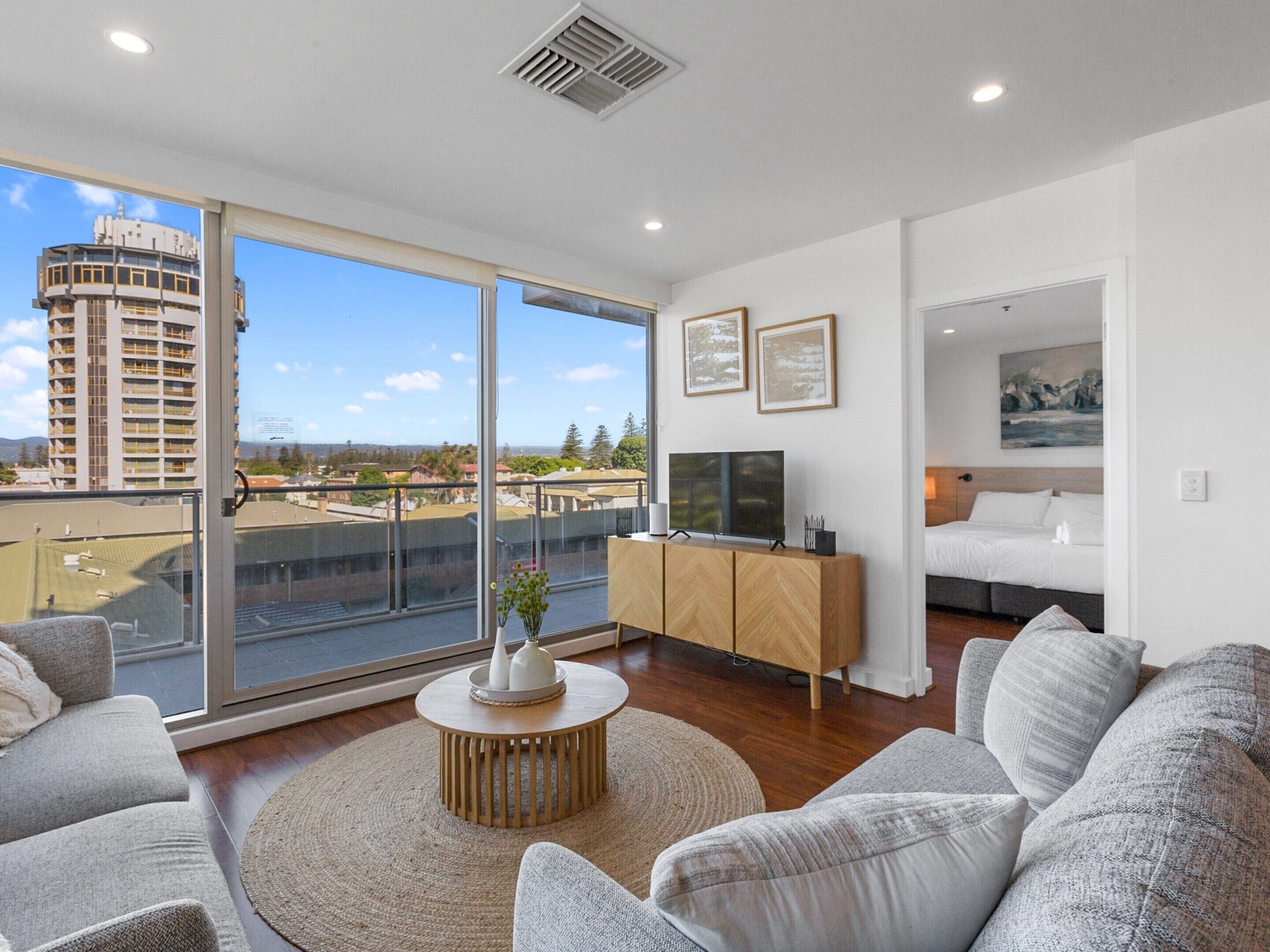 A modern living room with a large sliding glass door to a balcony, a flat-screen TV, a wooden cabinet, and a bed with a headboard.