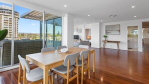 A modern living room with a wooden dining table, grey upholstered chairs, a sofa, and a large window offering a view of buildings and greenery. - Stylish 2-bed With Terrace, 3-min Walk to Beach (Adelaide)