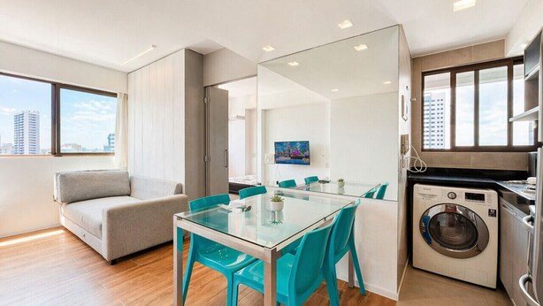 Eine moderne Wohnung mit einem Glastisch, blaugrünen Stühlen, einer Couch, einer Waschmaschine und Blick auf die Stadt.