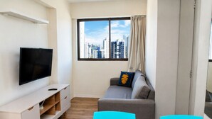 Ein modernes Wohnzimmer mit einem Flachbildfernseher, einem grauen Sofa, einem Glastisch und Blick auf die Stadt durch ein großes Fenster.