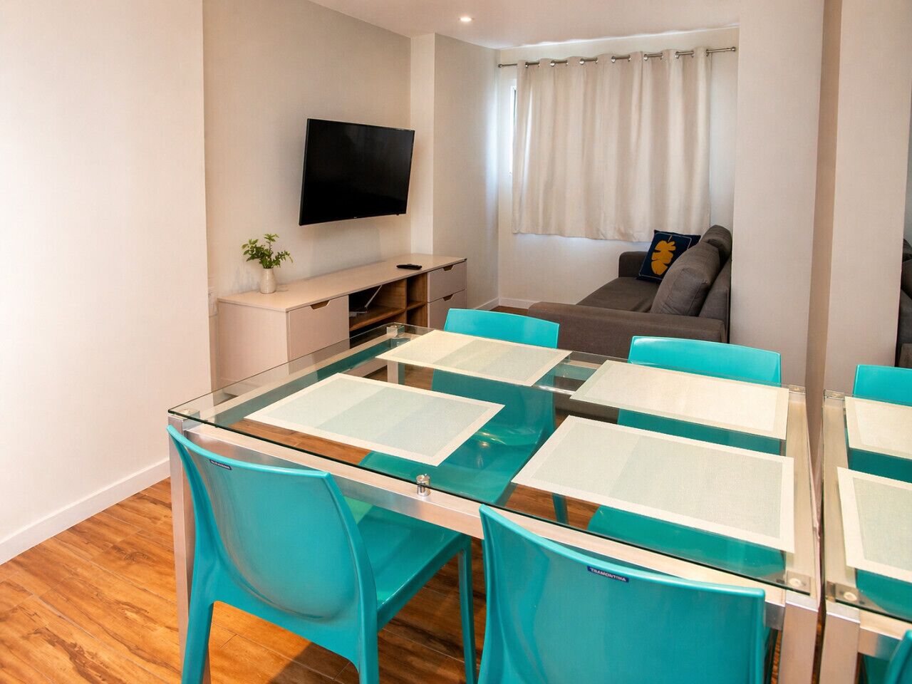 Een moderne kamer met een glazen eettafel, teal stoelen, een flatscreen televisie, een bank en een raam met gordijnen.
