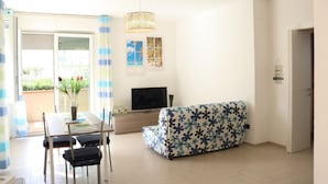 Ein Wohnzimmer mit einer Couch, einem Fernseher, einem Esstisch mit Stühlen und einem Balkon mit Vorhängen.