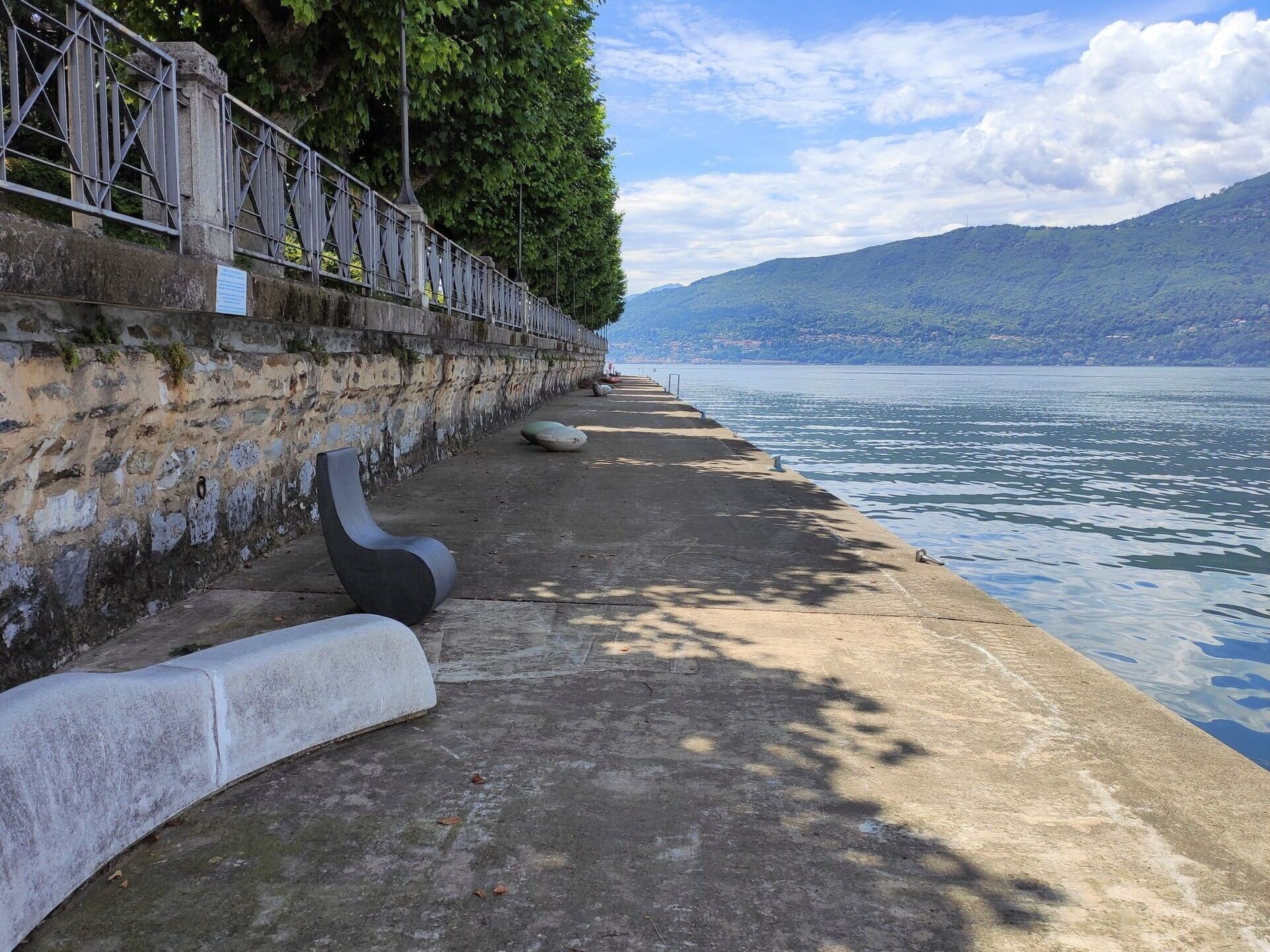 Una passeggiata sul lungolago in cemento, con un muro in pietra e una ringhiera metallica, affacciata su un lago tranquillo e montagne in lontananza.