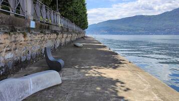 Una passeggiata sul lungolago in cemento, con un muro in pietra e una ringhiera metallica, affacciata su un lago tranquillo e montagne in lontananza.