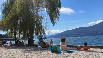 Una spiaggia di ciottoli con persone che si rilassano sotto un grande albero, affacciata su un lago con barche e montagne sullo sfondo.