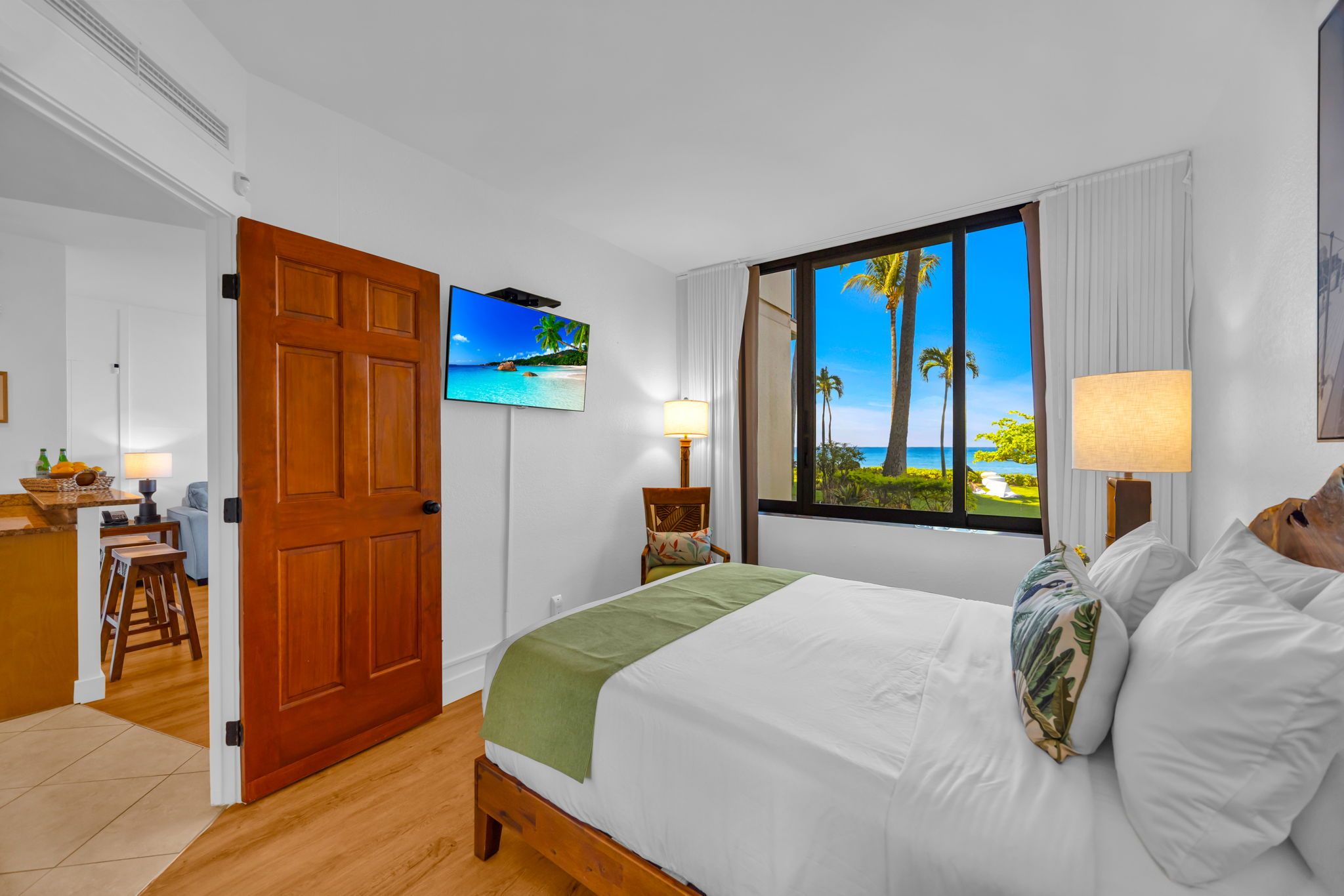 Ein Hotelzimmer mit einem hölzernen Bett, einem Fernseher, einem Fenster mit Blick auf Palmen und einen Strand sowie einem Schreibtisch mit Stuhl.