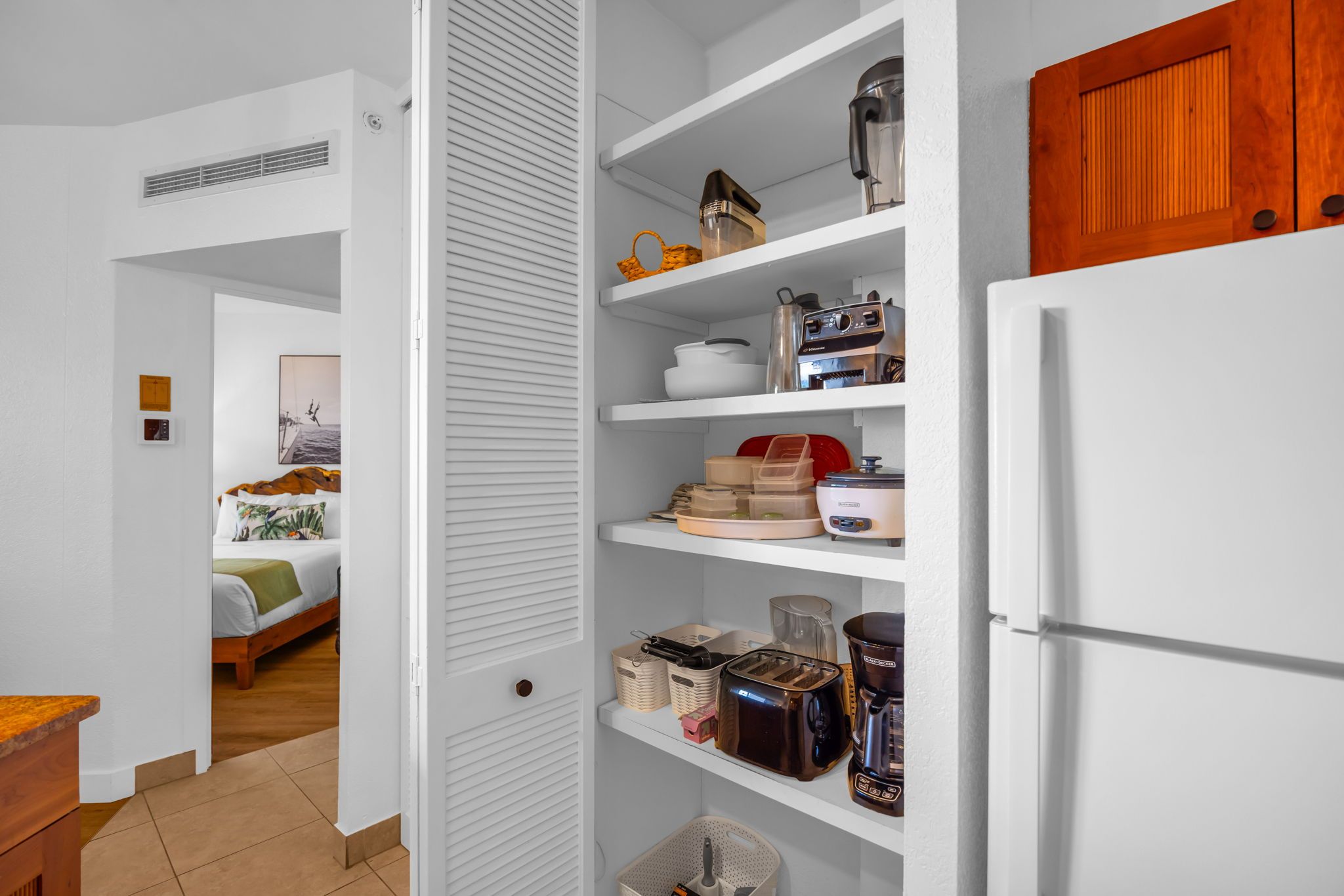 Ein Zimmer mit einem Bett, einem weißen Kleiderschrank und einer Küchenzeile mit Regalen, die Küchengeräte und Aufbewahrungsbehälter enthalten.