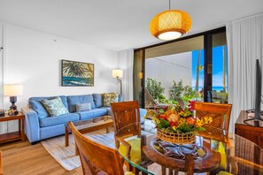 Ein Wohnzimmer mit Sofa, Sesseln, einem Glastisch, einer Lampe, einem Gemälde und Blick ins Freie.