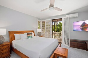 Un dormitorio con cama, mesitas de noche, ventilador de techo, televisor y balcón con contraventanas.