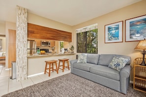 A living room with a grey sofa, two wooden stools, a coffee table, a lamp, and a kitchen area visible through a window. - 2 Grand Champions | Treeline View | Sleeps 10 | Free Car w/ 6+ Nts | ML-3777 (Wailea)