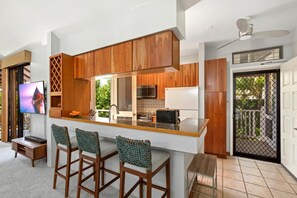 A modern kitchen with wooden cabinets, a built-in island with bar stools, a flat-screen TV, and a view of greenery outside. - Grand Champions | Ocean View 4 BR, Sleeps 10 | Car Incl. w/ 6+ Nights | GCH ML-3780 by KBM (Wailea)