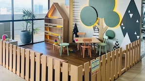 Ein Kinderspielbereich mit einem hölzernen Spielhäuschen, Sitzgelegenheiten und einer Wand mit geometrischen Mustern.