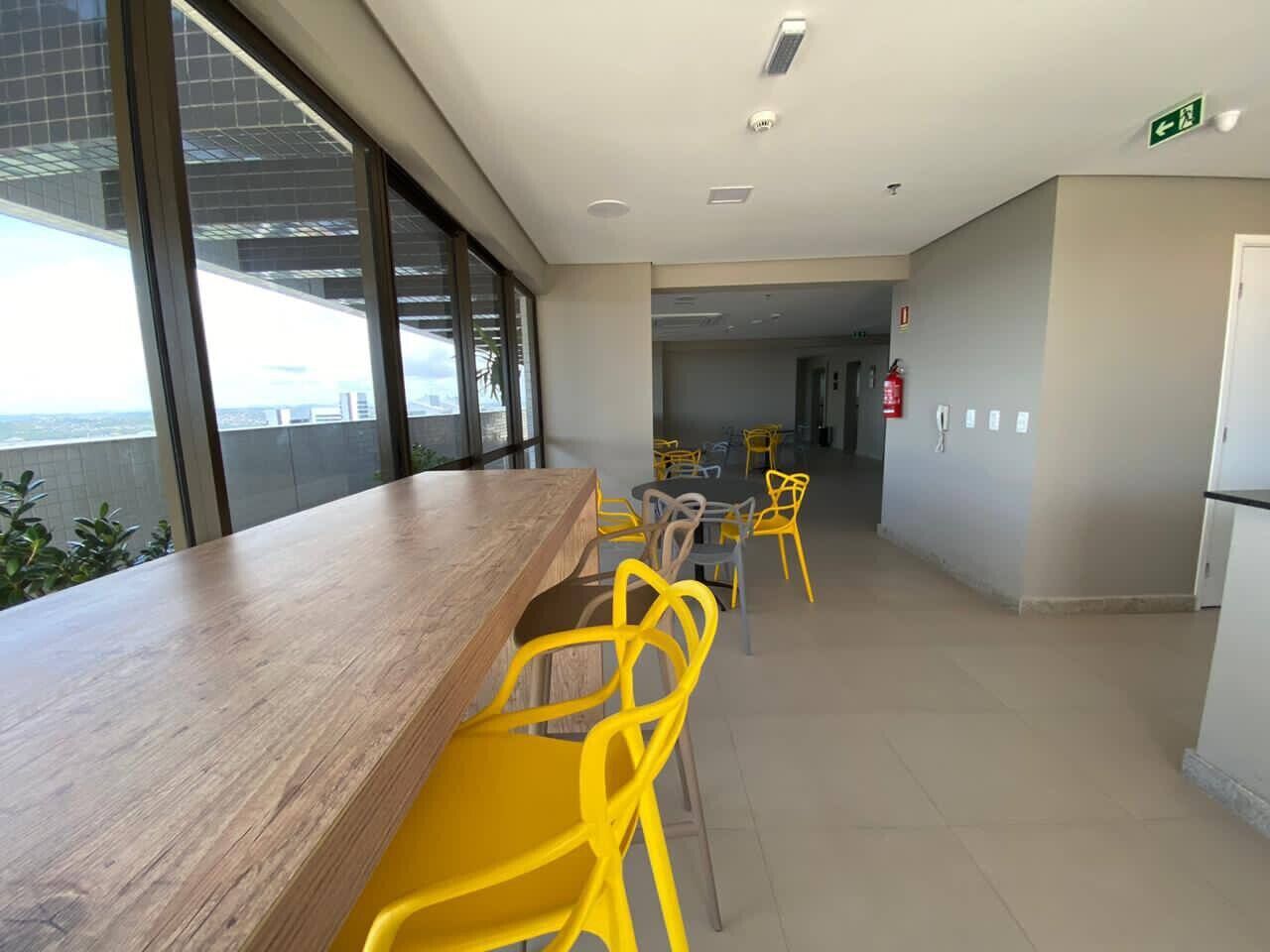 A modern indoor space with a long wooden table and yellow chairs, large windows, and a visible exit sign.