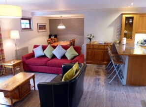 Een woonkamer met een rode bank, een zwarte fauteuil, een houten eettafel en een keuken met een bar.