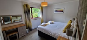 Een slaapkamer met een bed, nachtkastje, spiegel, raam met gordijnen en een aan de muur hangend schilderij.
