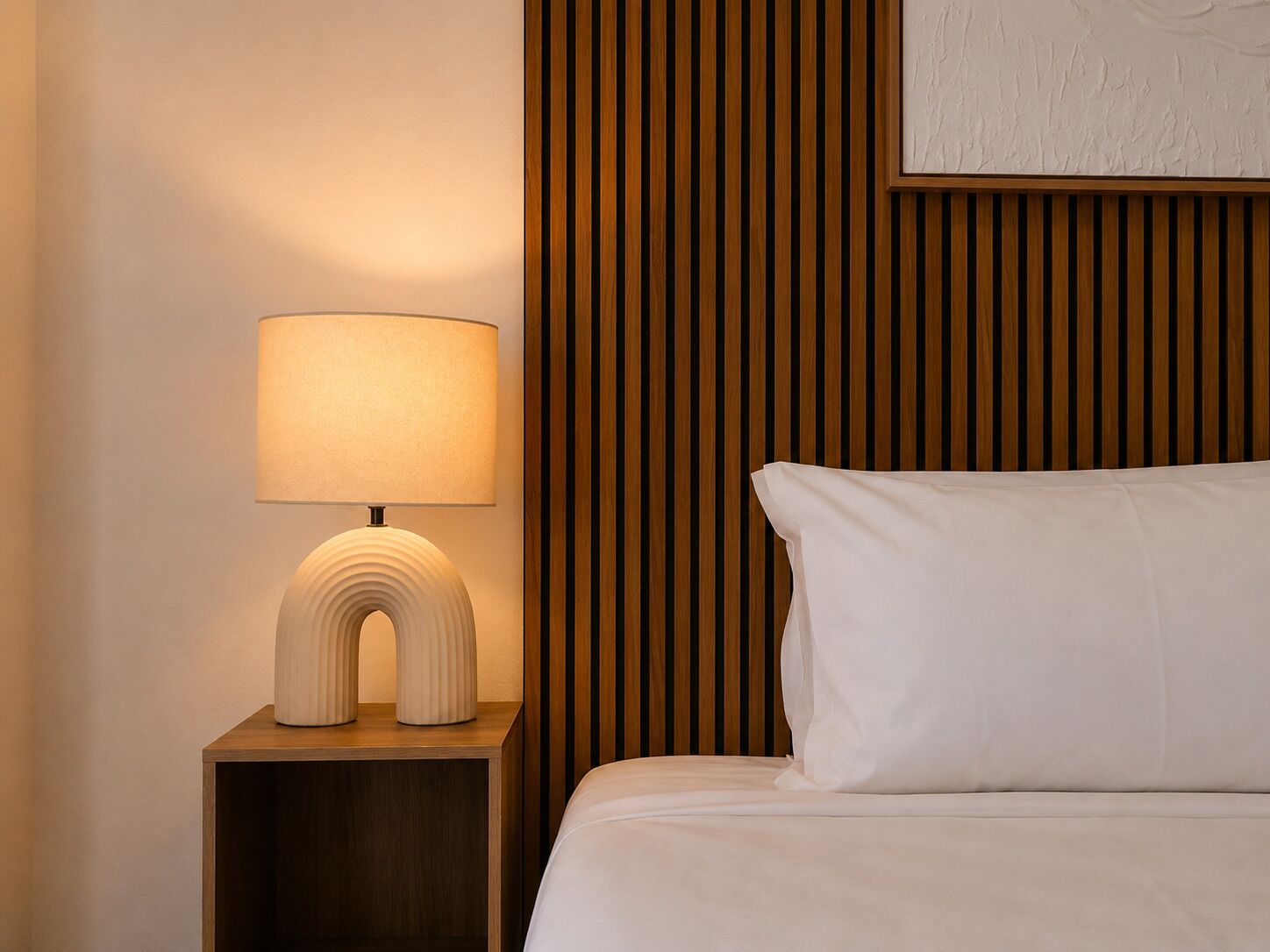 Habitación de hotel ordenada con lámpara de noche, cama con ropa blanca y pared de paneles de madera.