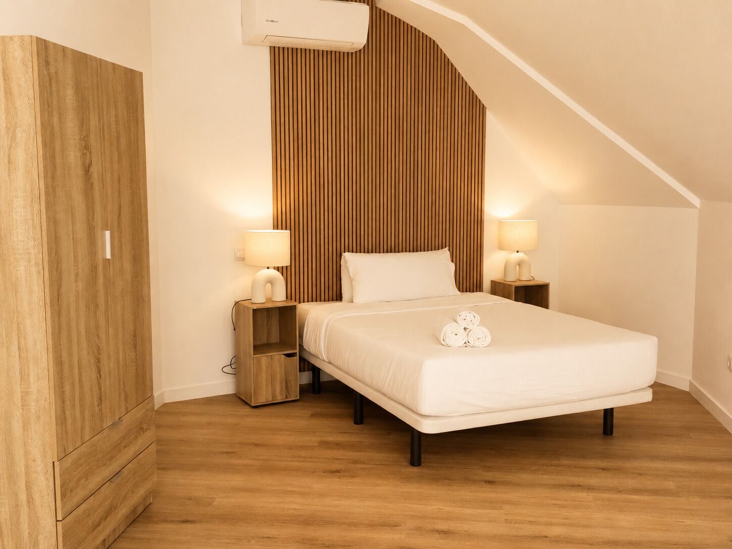 Camera da letto con soffitto spiovente, un letto con lenzuola bianche, due lampade da comodino e un'armadio in legno.