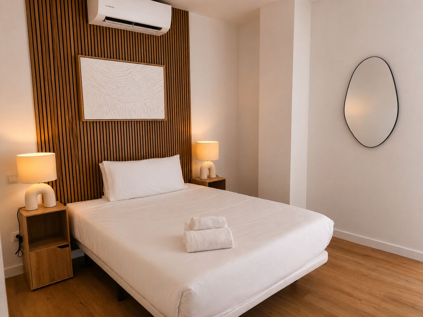 A hotel room with a bed, two bedside lamps, a wall-mounted mirror, and an air conditioning unit.