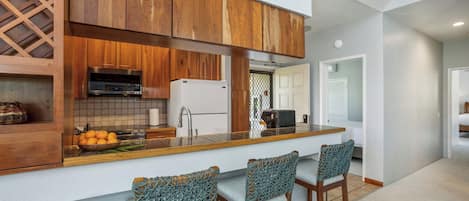 Una cocina moderna con muebles de madera, horno empotrado y pileta de acero inoxidable. Hay tres taburetes de bar con asientos de mimbre.