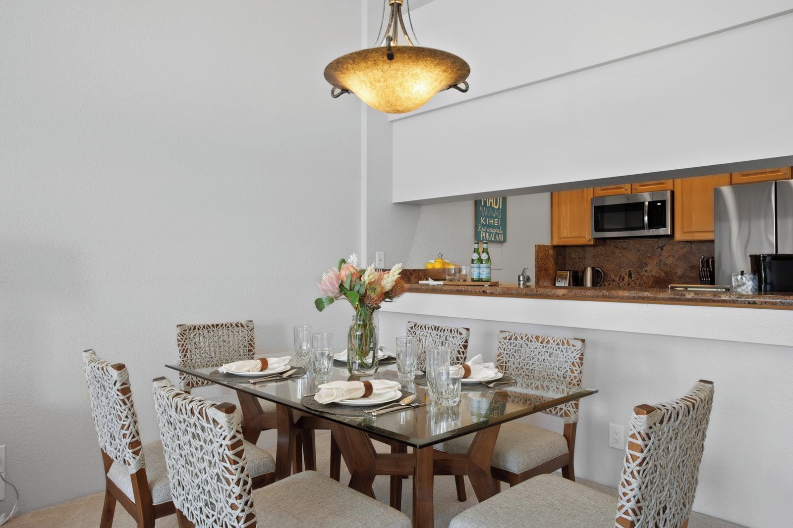 A dining area with a glass table, woven chairs, a vase with flowers, and a kitchen in the background.