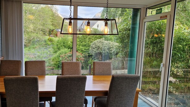 Een eetruimte met een tafel en stoelen, een hanglamp en uitzicht op groen door grote ramen.