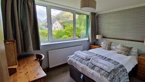 Een slaapkamer met een groot bed, een houten bureau, een radiator en een raam met uitzicht op groen en een gebouw.