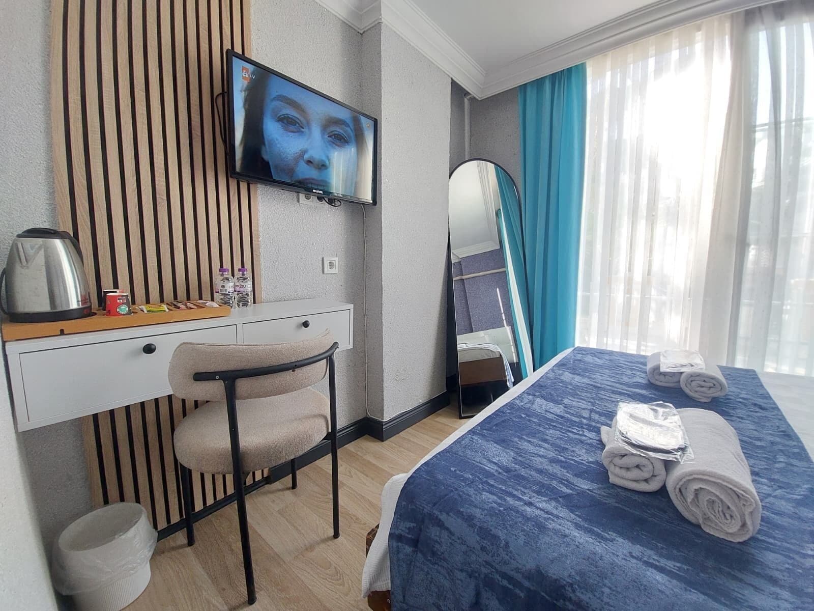 A hotel room with a bed, a desk with a chair, a television, a mirror, and a window with curtains.