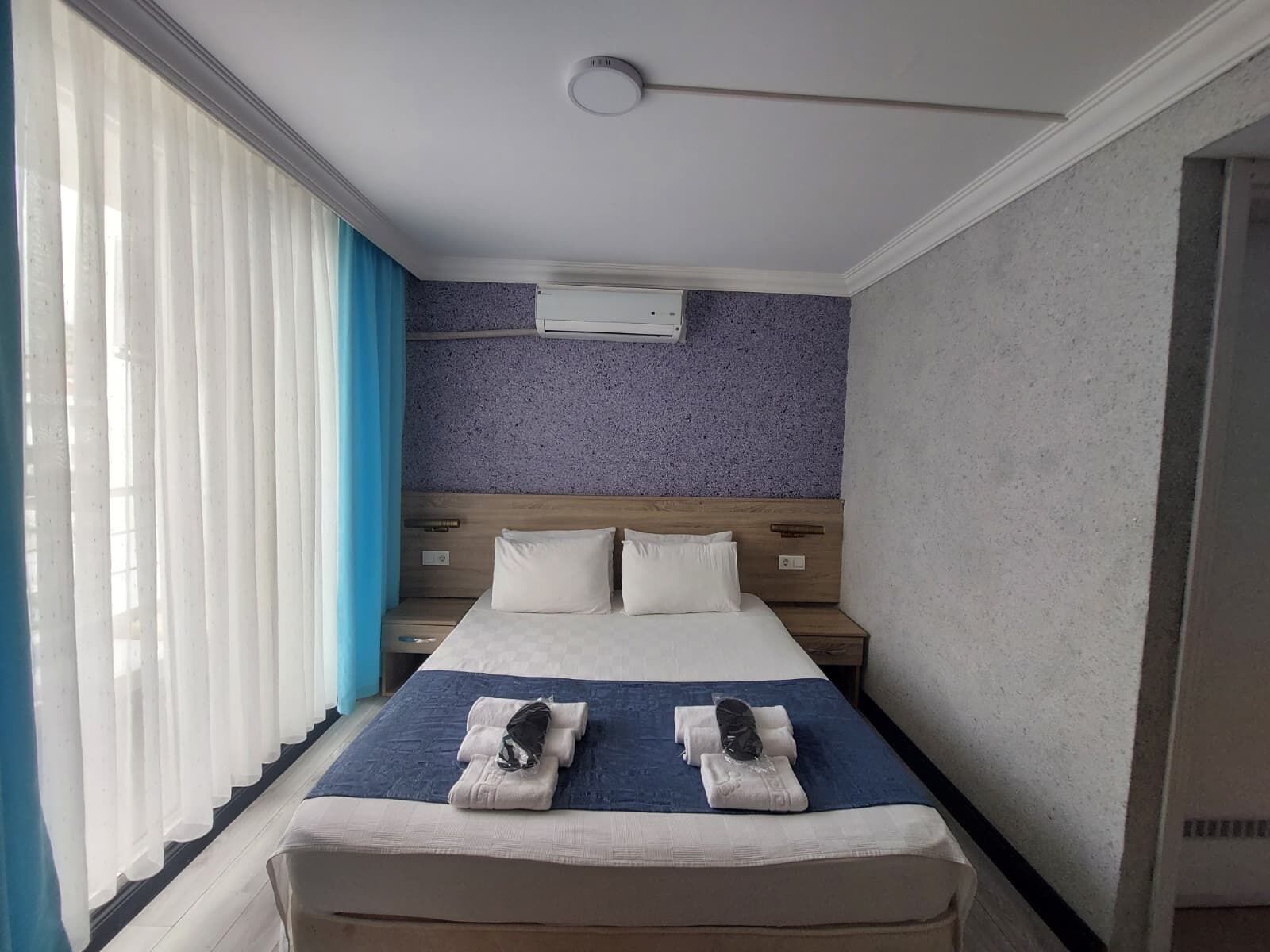 A hotel room with a bed, a headboard, a window with curtains, and a wall-mounted air conditioning unit.