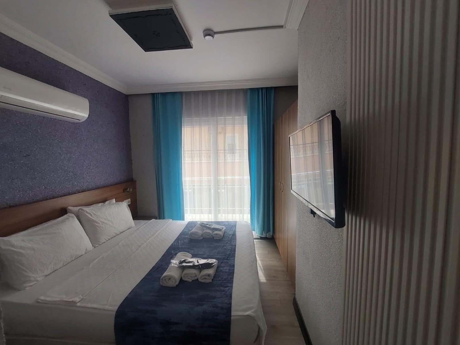 Una habitación de hotel con cama, televisión, ventana con cortinas y un respiradero en el techo.