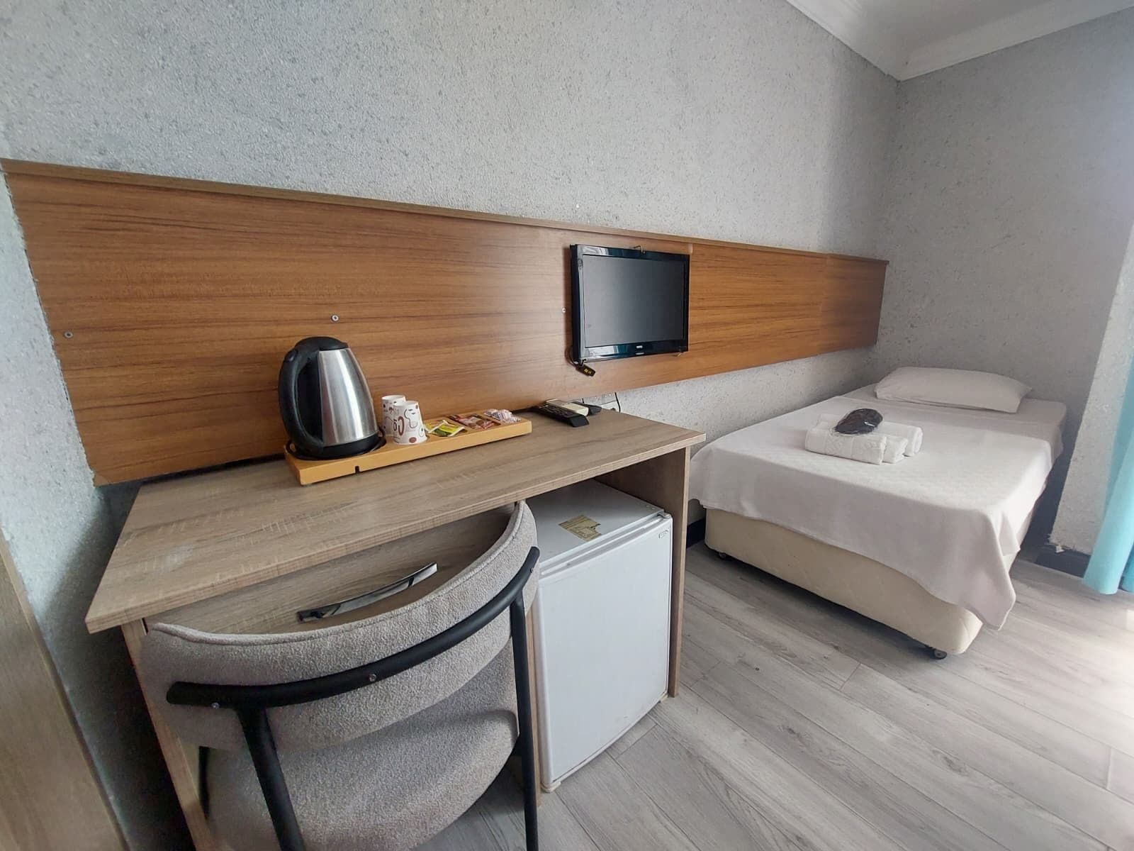 Habitación de hotel compacta con escritorio de madera, refrigerador pequeño, cama con ropa de cama blanca y televisor montado en la pared.