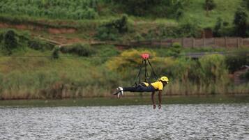 Une personne suspendue en l’air par un câble, portant un casque jaune et une tenue noire, au-dessus d’une étendue d’eau, avec une végétation verdoyante en arrière-plan.