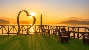 A deck with a heart-shaped sculpture, a table, chairs, and a view of the sunset over water.
