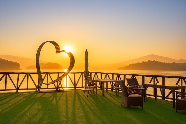 Une terrasse agrémentée d’une sculpture en forme de cœur, d’une table, de chaises et offrant une vue sur le coucher de soleil sur l’eau.