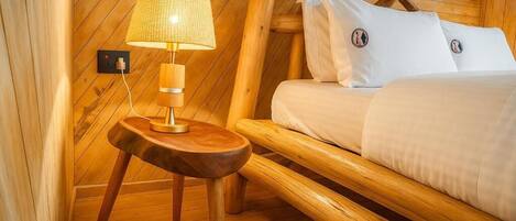 Una cama de madera con ropa de cama blanca, una mesita de noche de madera con una lámpara, y un interruptor fijado a la pared.
