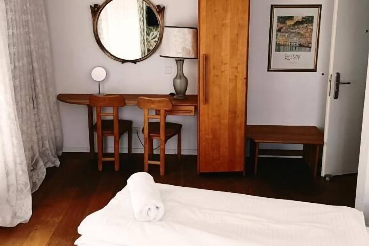 A hotel room with a bed, a desk with two chairs, a wooden wardrobe, a round mirror, a lamp, and a door.