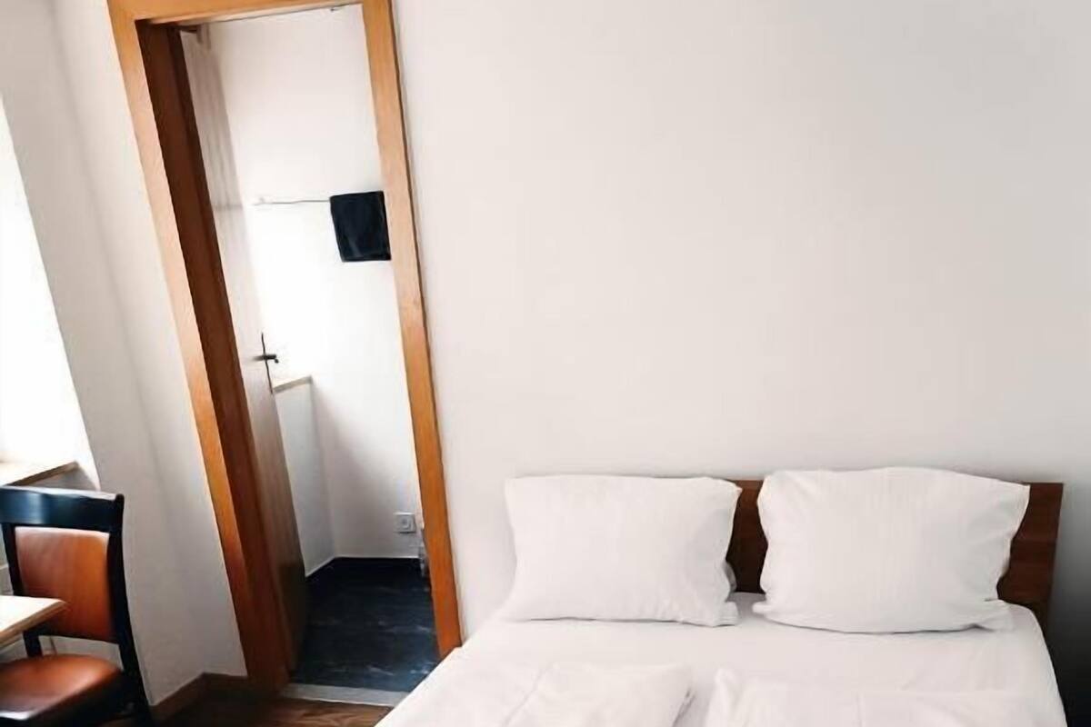 A single bed with white linens, a wooden headboard, and a chair to the left.