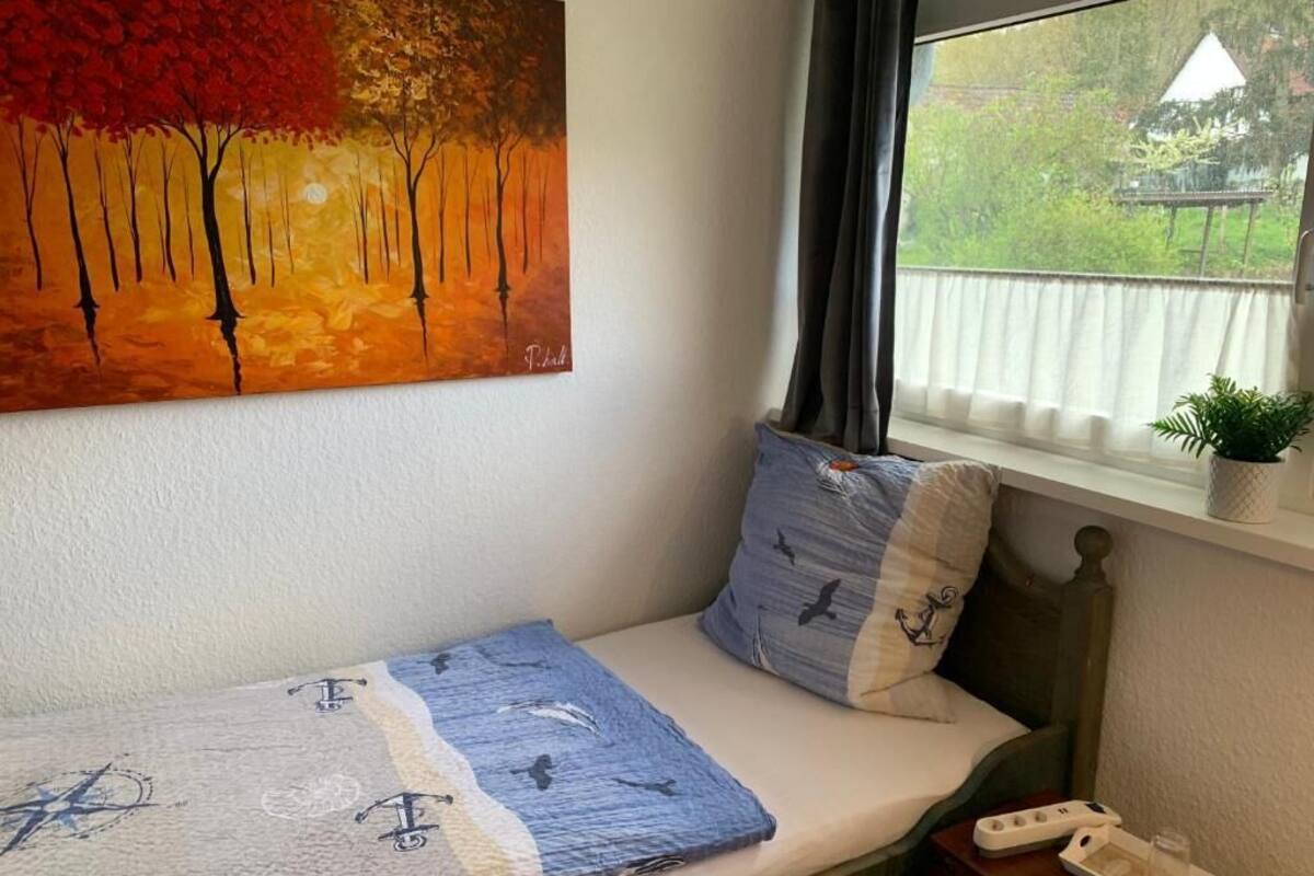 A room with a bed, a painting, a window with curtains, a plant, and a bedside table.