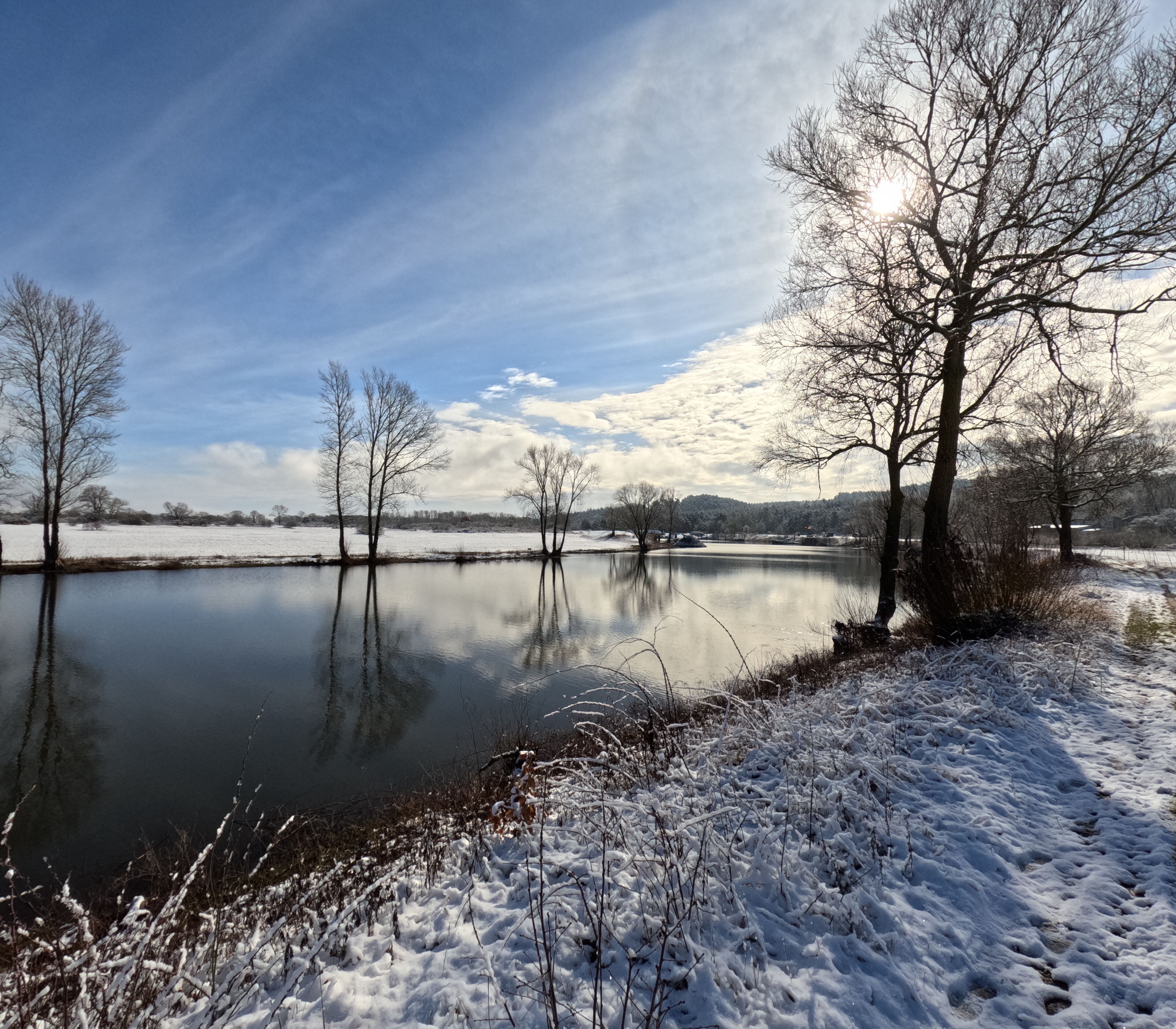 A serene winter landscape with a calm lake, snow-covered ground, and leafless trees.
