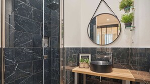 Un baño con baldosas de mármol negro, un espejo redondo, un tocador de madera y una planta en maceta.