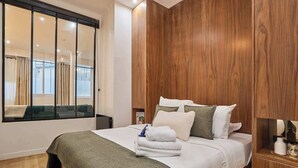 Un dormitorio moderno con cabecera de madera, cama con ropa blanca y varios almohadones, mesita de noche con lámpara, y vista a través de un ventanal.