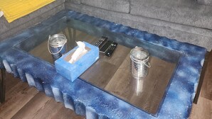 Un salon avec un canapé gris, des coussins à motifs, un plaid jaune, une table basse en verre avec des télécommandes et une boîte de mouchoirs, et un tableau accroché au mur.