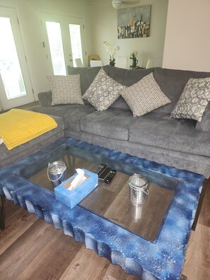 Ein Wohnzimmer mit grauem Sofa, gemusterten Kissen, einer gelben Decke, einem Glastisch mit Fernbedienungen und einer Taschentuchbox sowie einem Bild an der Wand.