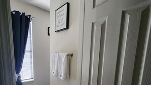 Un couloir avec une porte blanche, un panneau encadré accroché au mur, un porte-serviettes et un paillasson à carreaux.