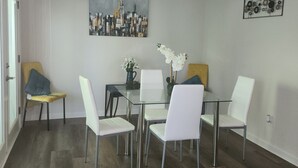 Un coin repas avec une table en verre, des chaises blanches, un tableau représentant un paysage urbain et un ventilateur de plafond.