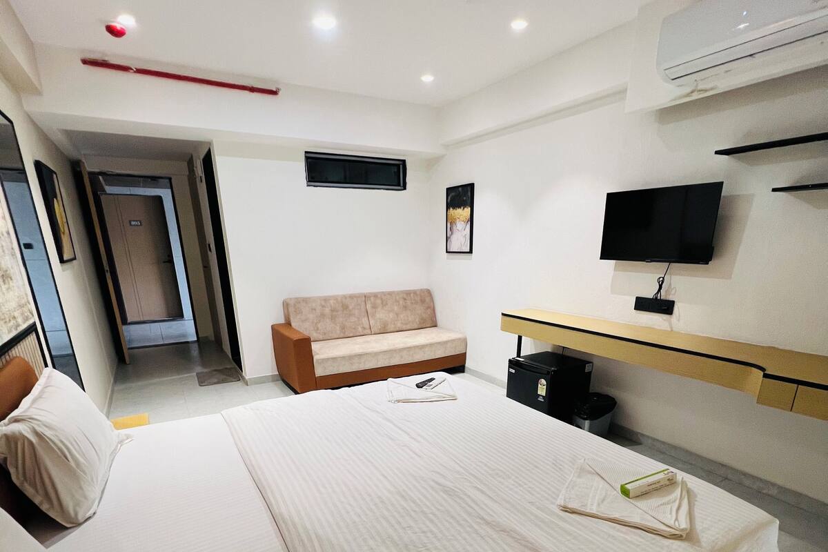 A hotel room with a bed, a sofa, a TV, and an air conditioning unit.
