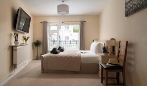 A bedroom with a bed, a bedside table with a lamp, a chair, a radiator, a TV, and a window with curtains.