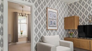 Un salon avec un papier peint à motifs, un canapé beige, un meuble en bois, un téléviseur et un tableau encadré accroché au mur.