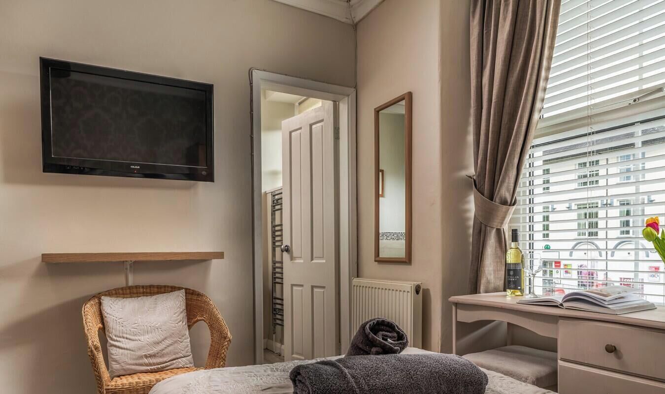 Hotellihuone, jossa on sänky, televisio, työpöytä tuolilla, peili ja ikkuna, josta on näkymä rakennuksiin.