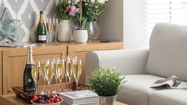 Un salon avec un meuble en bois, un canapé, une table basse sur laquelle se trouve un livre, un saladier rempli de fruits et une bouteille de champagne.