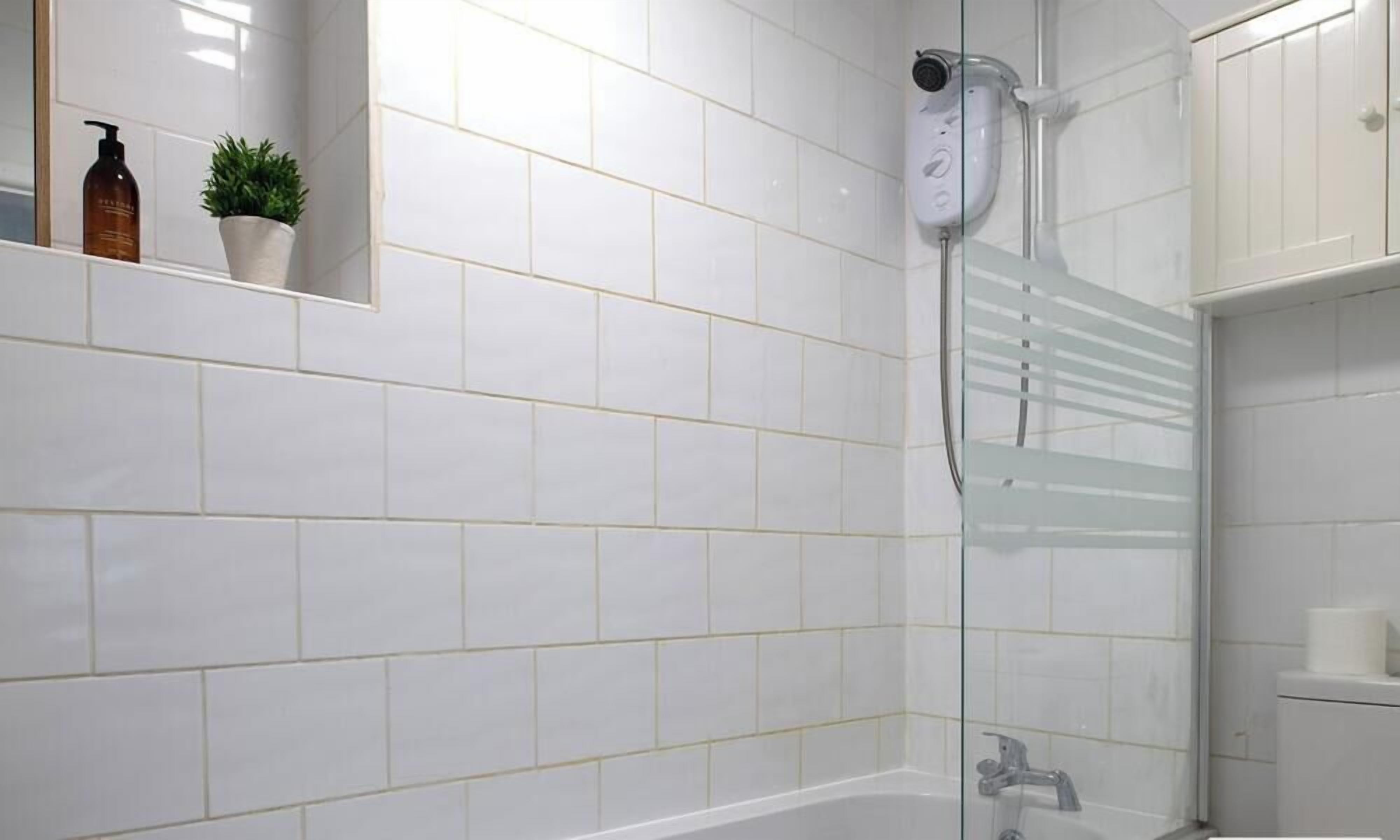 Een moderne badkamer met witte tegels, een douchecabine van glas, een handdouche en een klein plankje met toiletartikelen.